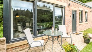 Un bâtiment moderne doté de grandes fenêtres en verre, d’un patio avec une table et des chaises, et d’une façade en briques.