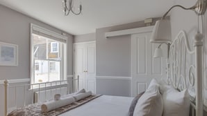 A neatly made bed with a floral-patterned blanket, a white headboard, and a window with a view of buildings outside. - Harbourside Cottage (Weymouth)