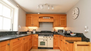 A kitchen with wooden cabinets, a stove, a microwave, and a clock on the wall. - Harbourside Cottage (Weymouth)
