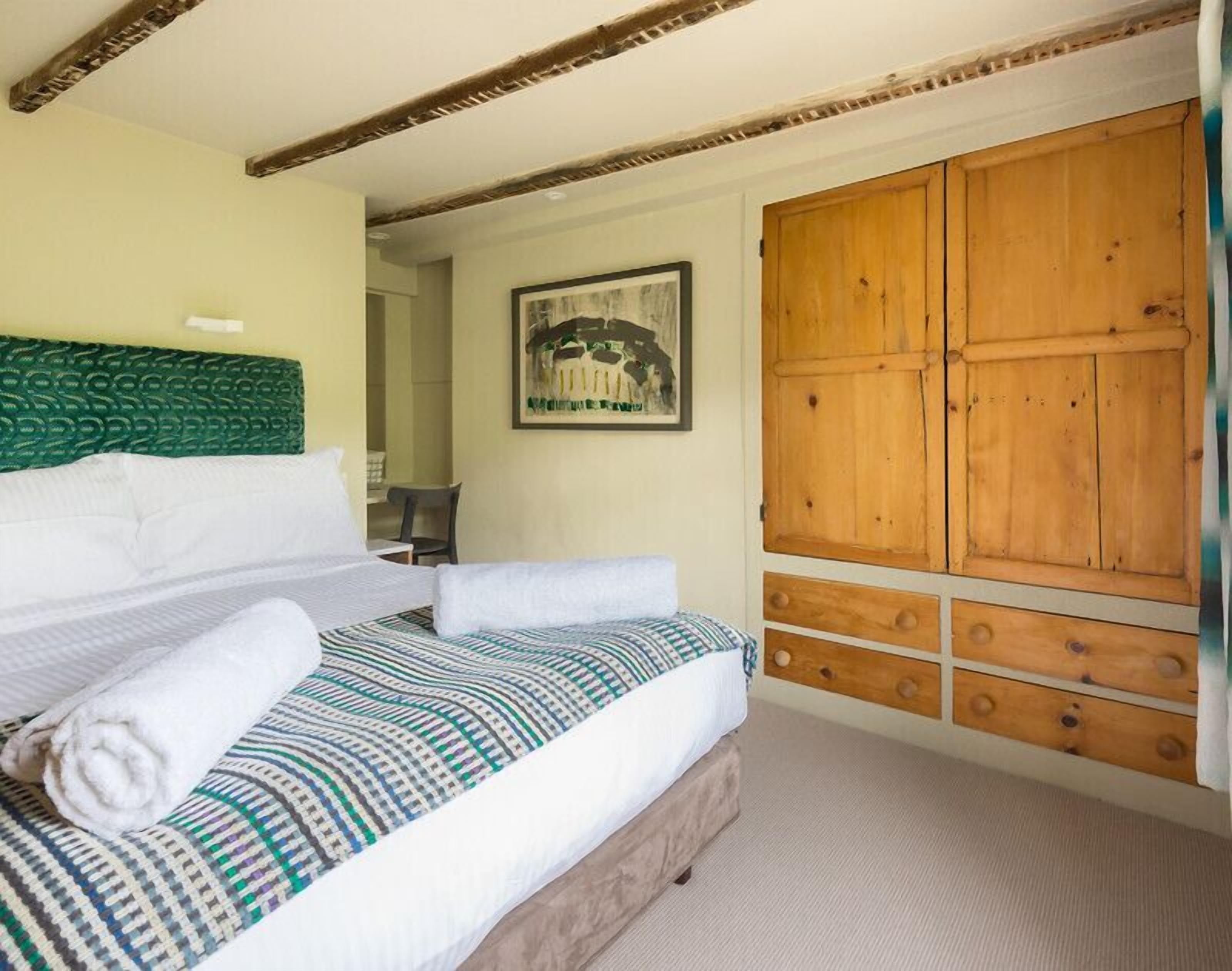 Un letto rifatto con cura, con una coperta a motivi decorativi e asciugamani bianchi arrotolati, un armadio in legno con diversi cassetti e un quadro appeso al muro.