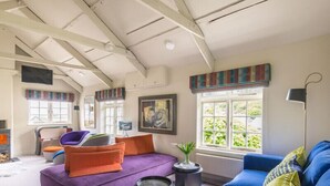Ein geräumiger Raum mit hoher Decke, einem großen Fenster mit Blick auf Grünflächen, einem blauen Sofa, einem violetten Sofa und einem Sessel.