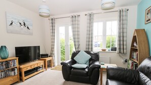 A living room with a black leather sofa, a wooden coffee table, a television on a wooden stand, a bookshelf, and a window with curtains. - Puffin Cottage (Filey)
