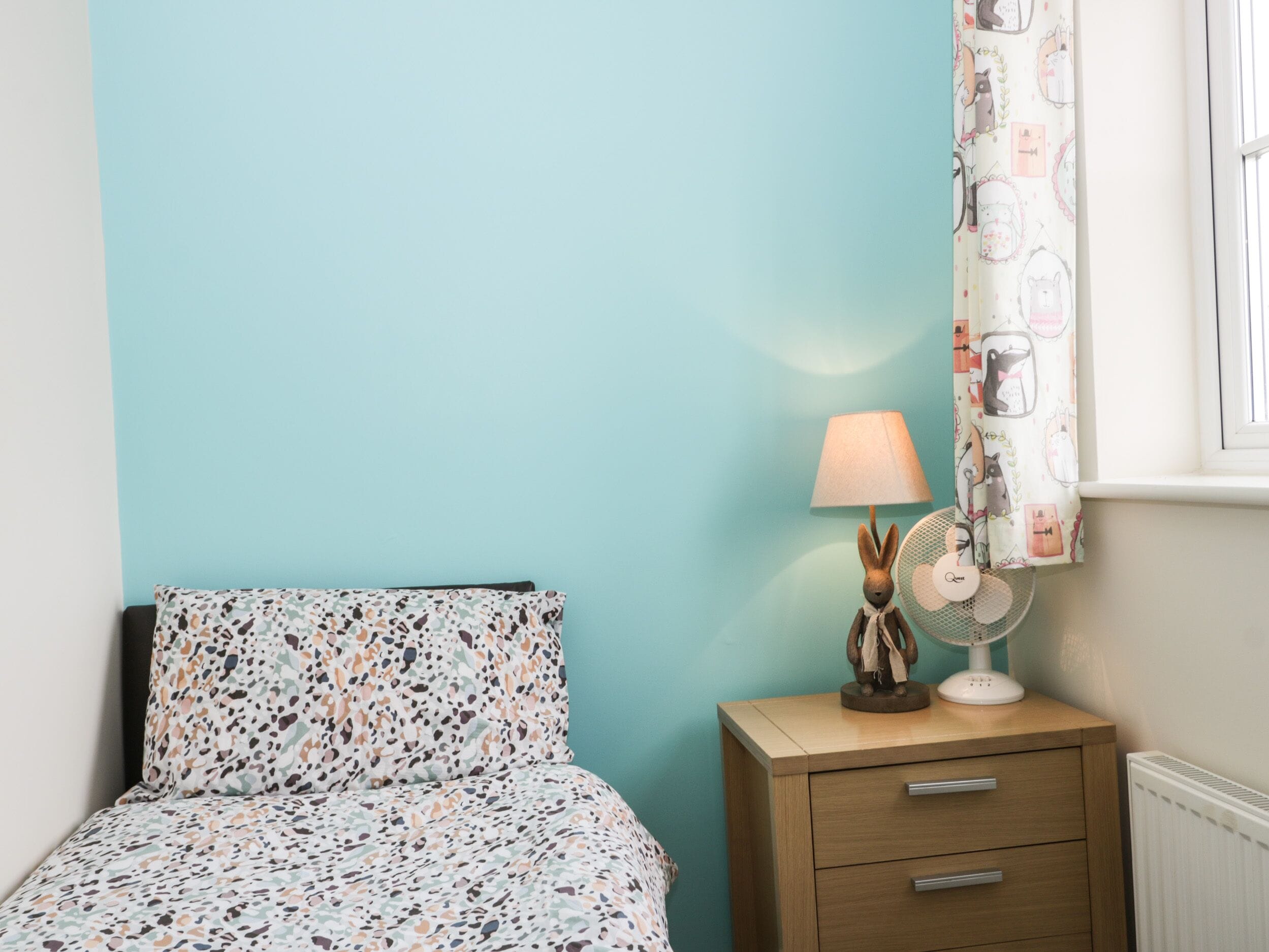 A bedroom with a bed, a bedside table with a lamp and a fan, and a window with curtains.
