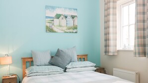 A bedroom with a bed, a bedside table, a lamp, a radiator, and a window with curtains. - Puffin Cottage (Filey)