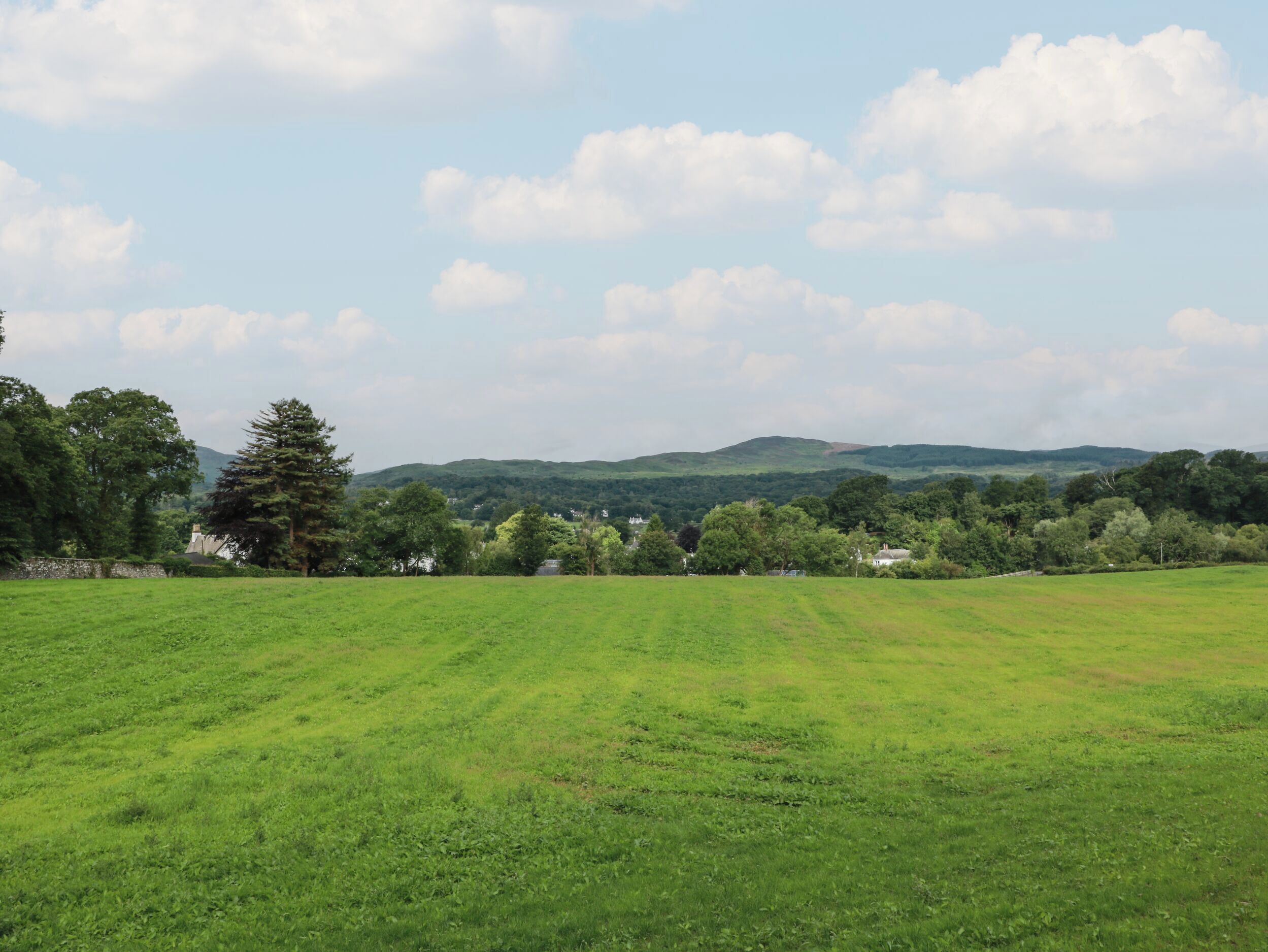 A wide, open grassy field with a few trees and a distant hill range.