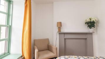 A room with a bed, a chair, a radiator, a window with a curtain, a lamp, and a plant on a shelf.