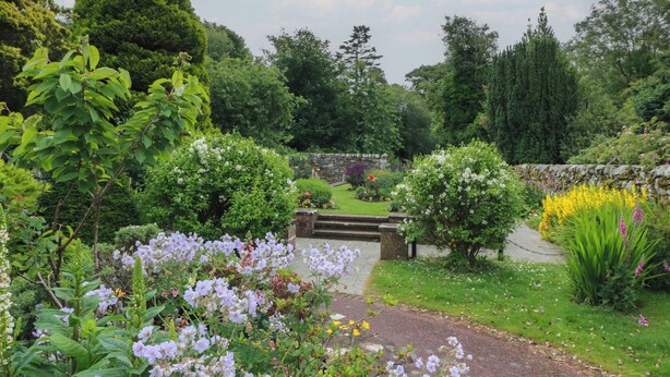 Ein Garten mit einer Steinbrücke, üppigem Grün und verschiedenen blühenden Pflanzen.