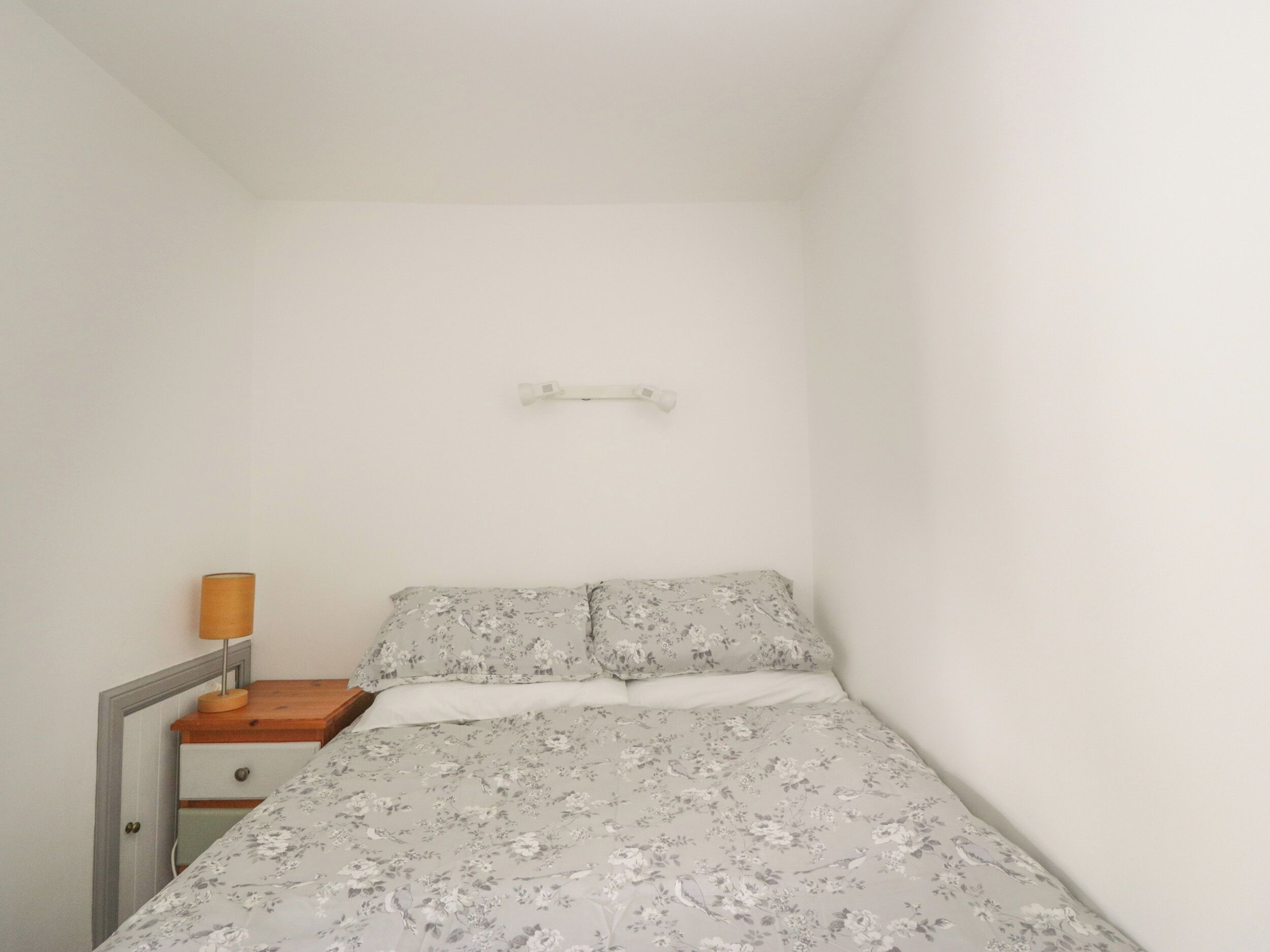 A small, single-bed room with a wooden nightstand, a lamp, and a wall-mounted shelf.
