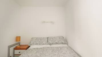 A small, single-bed room with a wooden nightstand, a lamp, and a wall-mounted shelf.