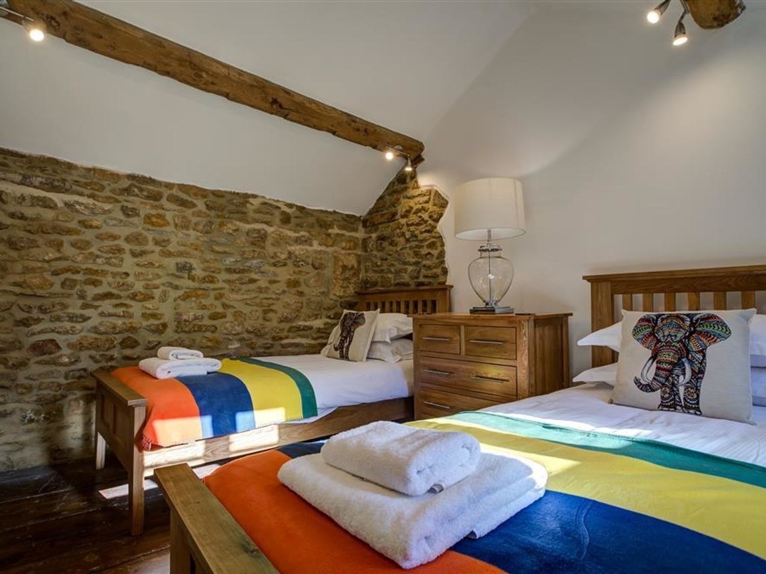 A cozy room with a stone wall, two beds, a wooden bench, and a lamp.