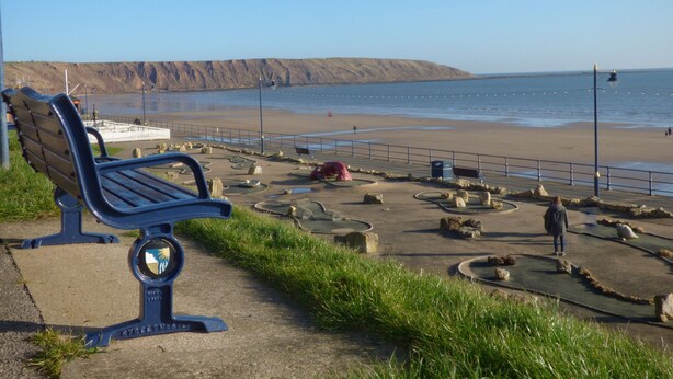 Un banc situé le long d’une promenade côtière, offrant une vue sur la plage et les falaises.
