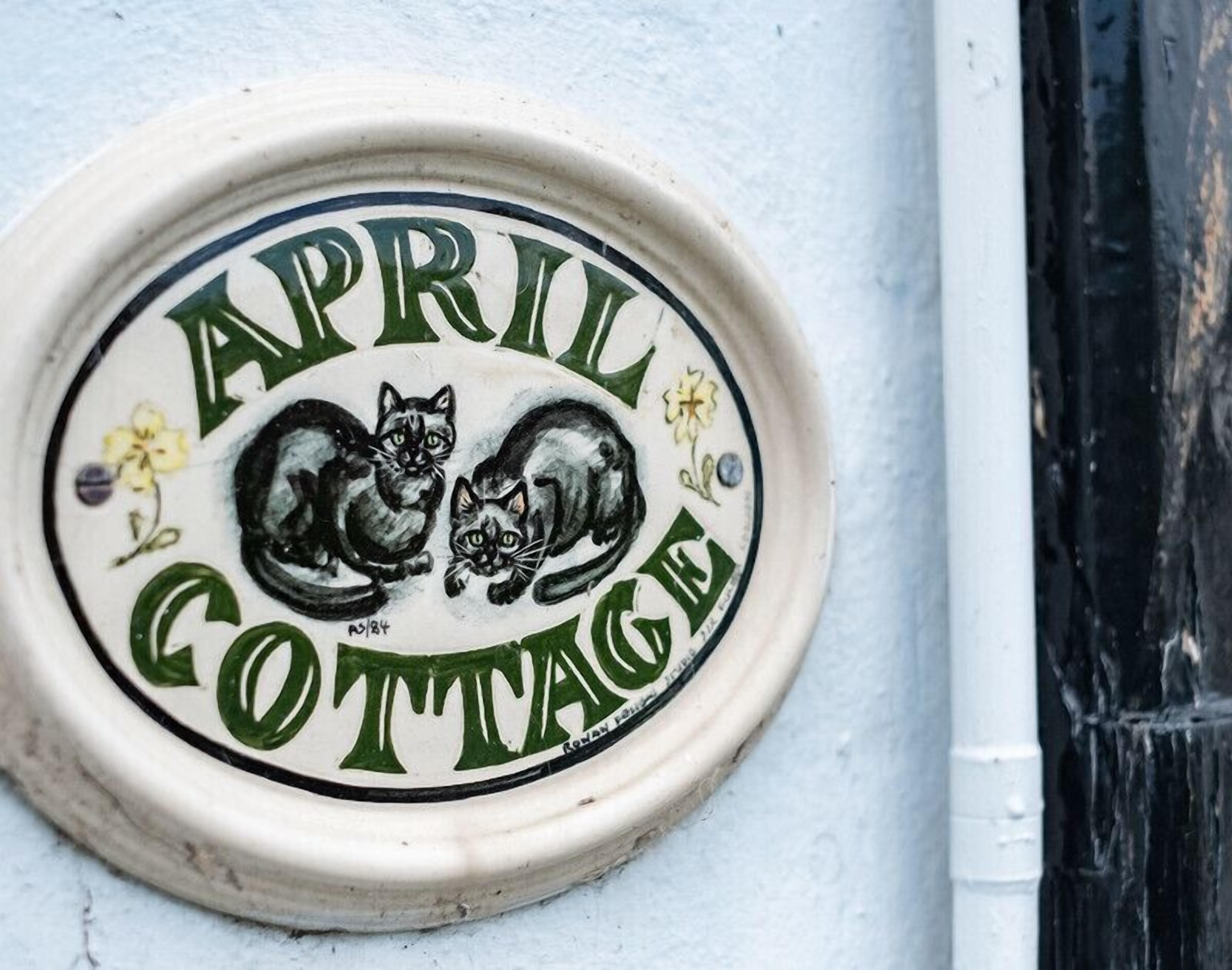 A ceramic plaque with the text "APRIL COTTAGE" and two black cats, one curled up and one standing, with a small flower on each side.