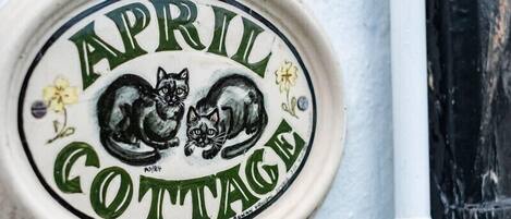A ceramic plaque with the text "APRIL COTTAGE" and two black cats, one curled up and one standing, with a small flower on each side.