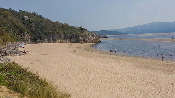 Une plage de sable où l’on peut se baigner et se promener, entourée de verdure et de collines.