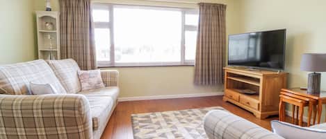 Una sala de estar con un sofá, un televisor sobre un mueble de madera, una lámpara y una ventana con cortinas.