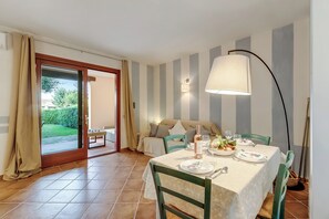 A dining area with a table set for four, a sofa, and a view of a garden through a sliding door. - Il Borgo 10/5 700m From The Beach (Loiri Porto San Paolo)