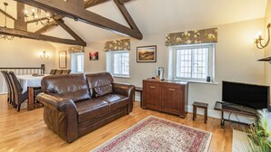 A living room with a brown leather sofa, wooden beams, a dining table with chairs, a TV, and a patterned rug. - Dales Croft Apartment (Buxton)