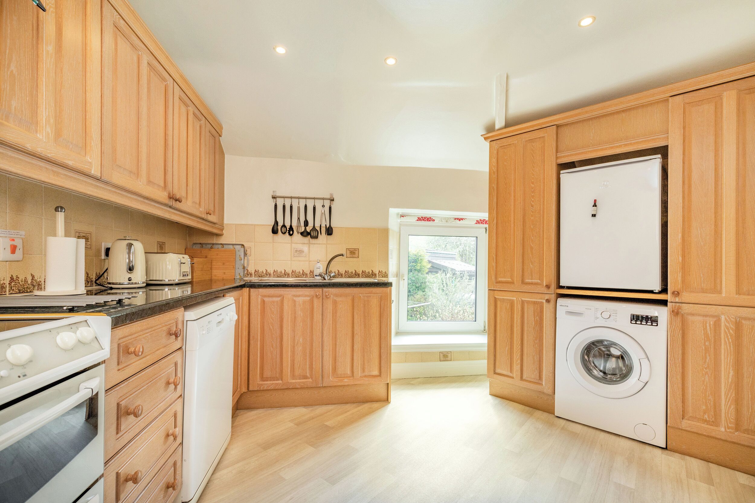 A modern kitchen with wooden cabinets, a built-in oven, a dishwasher, a washing machine, and a window providing natural light.