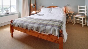 A bedroom with a wooden bed, a large window with curtains, a nightstand with a lamp, and a chair with a book. - Worthy House (Wadebridge)