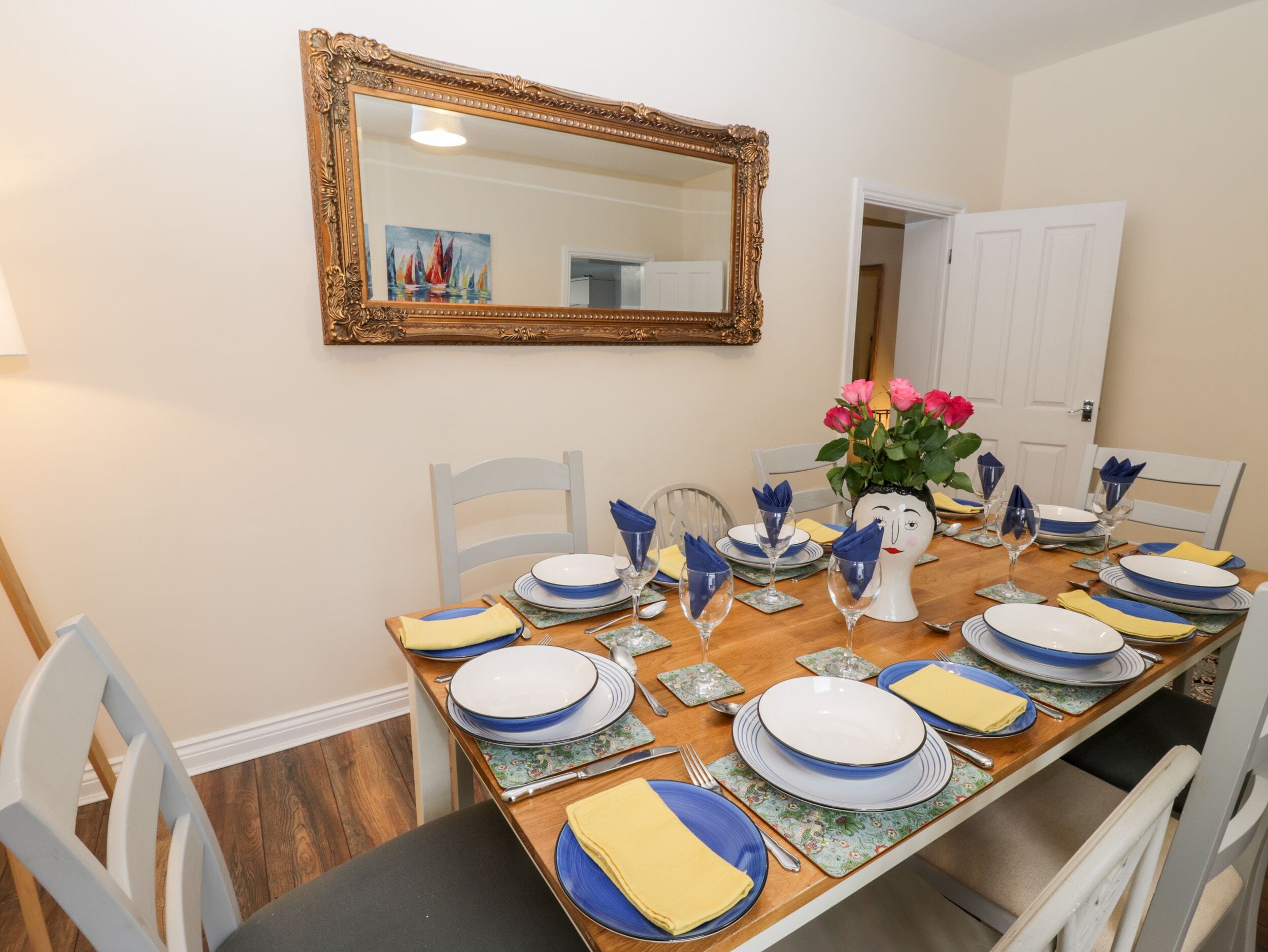 A dining room with a wooden table set for four, white chairs, a mirror on the wall, and a door in the background.