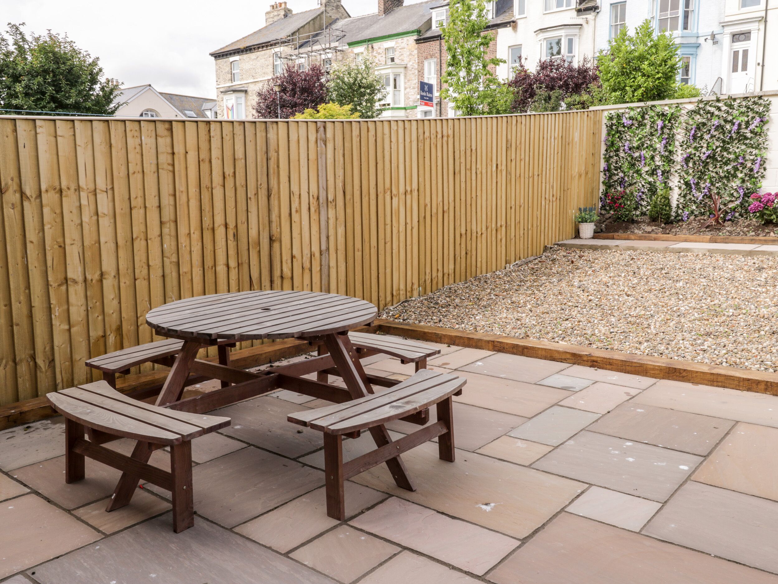 Una mesa de madera con bancos sobre un patio empedrado, cercado por una valla de madera y con un jardín de grava y plantas.