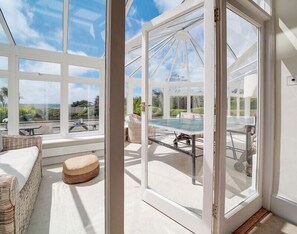 A conservatory with a glass table and chairs, a cushioned bench, and a view of the outdoors. - The Haven (Wadebridge)