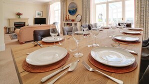A dining area with a wooden table set for a meal, surrounded by comfortable seating and large windows. - The Haven (Wadebridge)