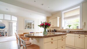 A modern kitchen with a central island, light-colored cabinetry, and a dining area with chairs. - The Haven (Wadebridge)