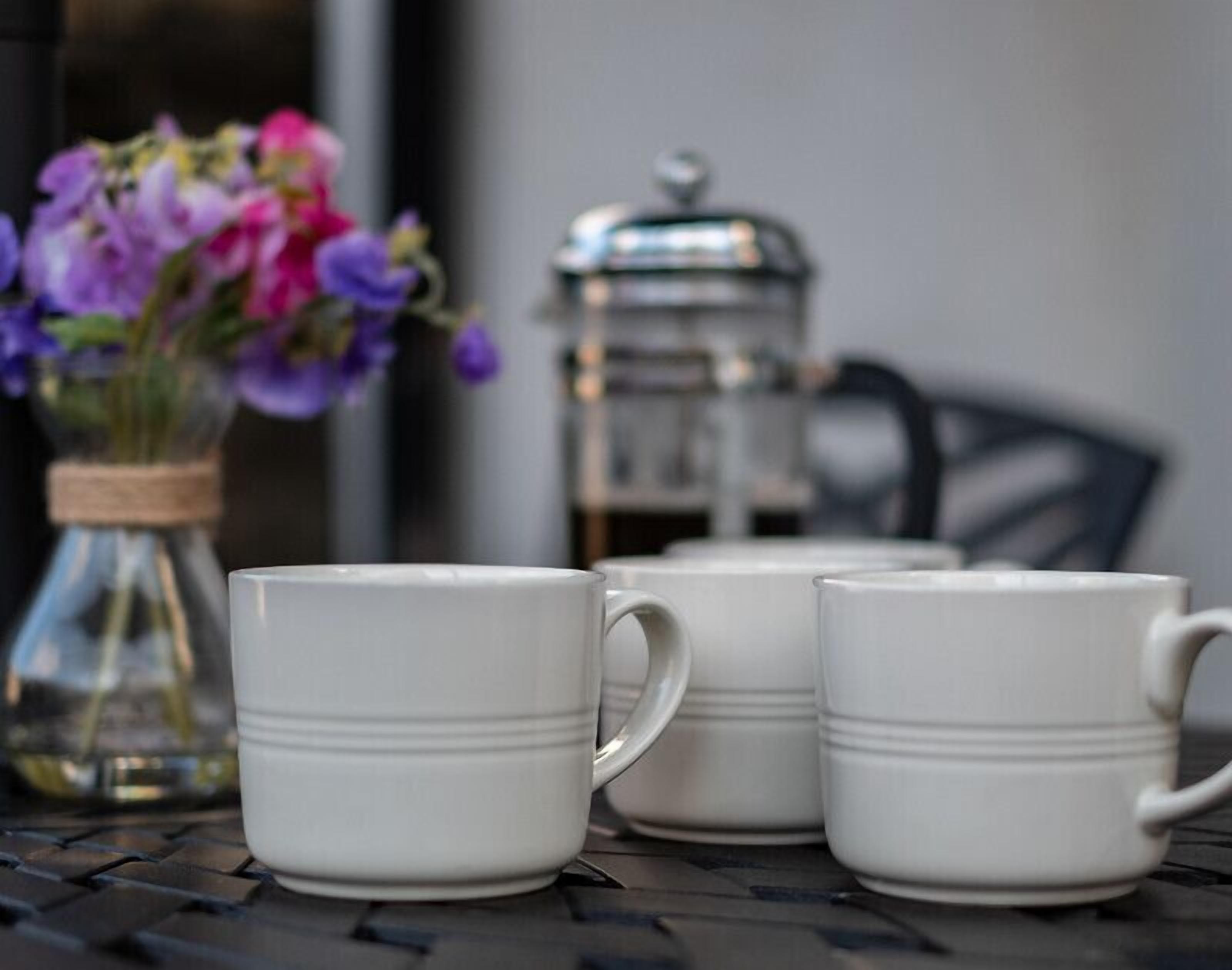 Three white ceramic mugs on a metal table with a blurred background featuring a metal container and a vase with purple flowers.