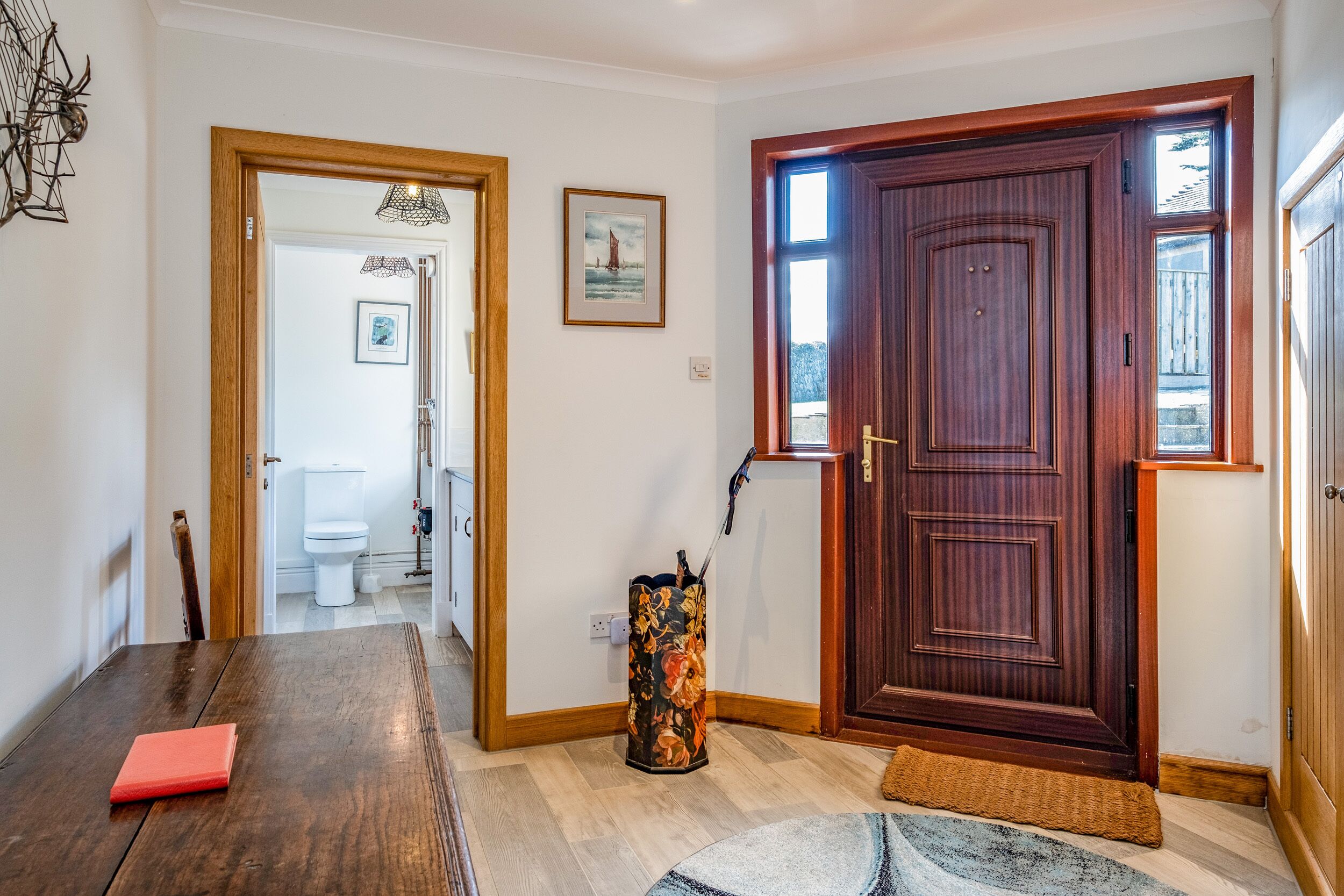 Una habitación con una puerta de madera, una mesa de comedor con un objeto rojo, un jarrón estampado y una alfombra.