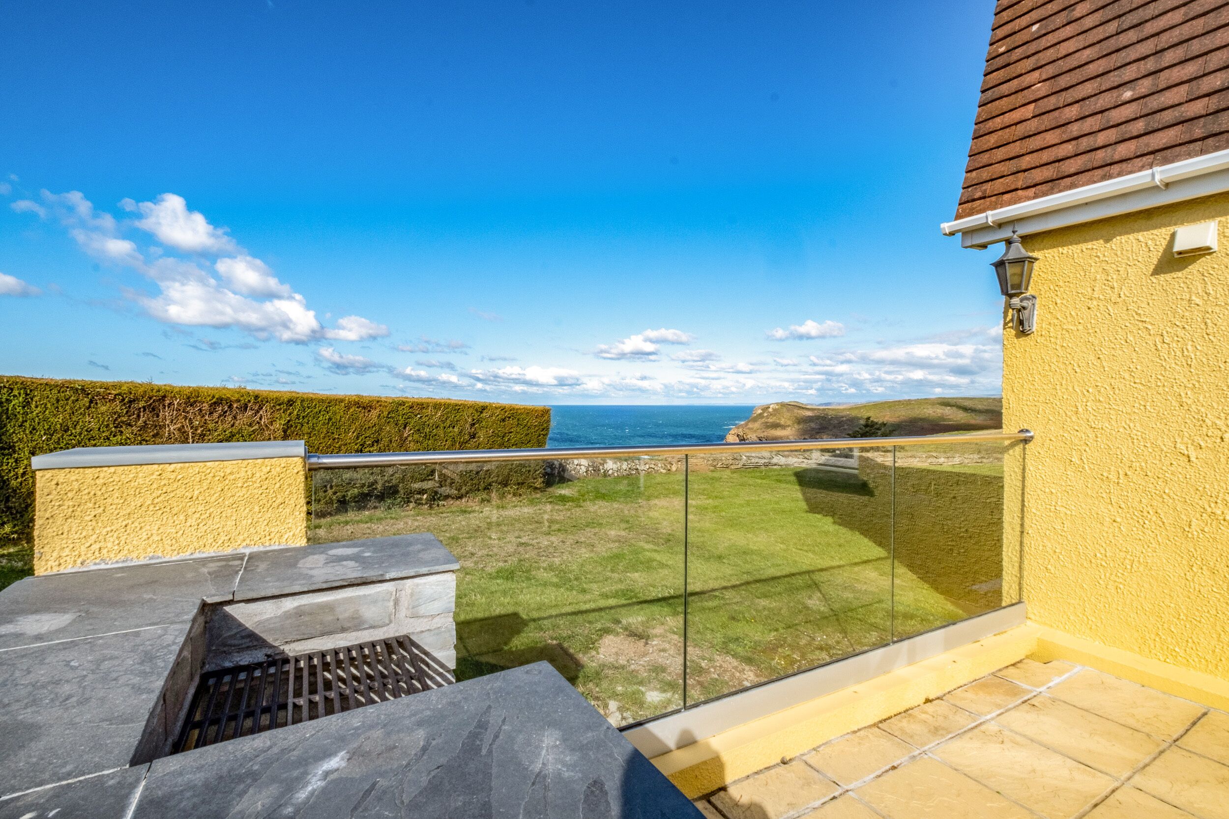 A balcony with a glass railing overlooking a grassy area and a distant view of the sea.