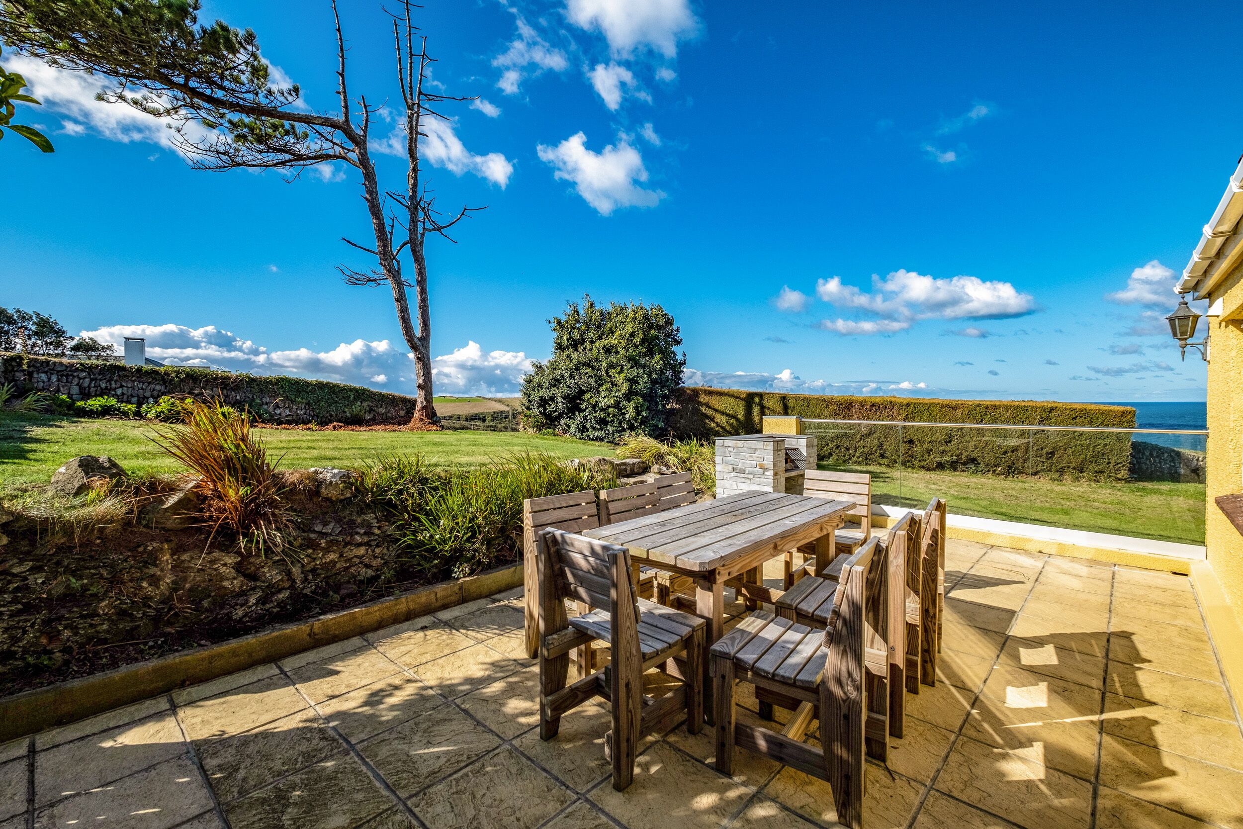 A patio with a wooden table and chairs, a stone wall, and a view of a grassy field and distant hills.