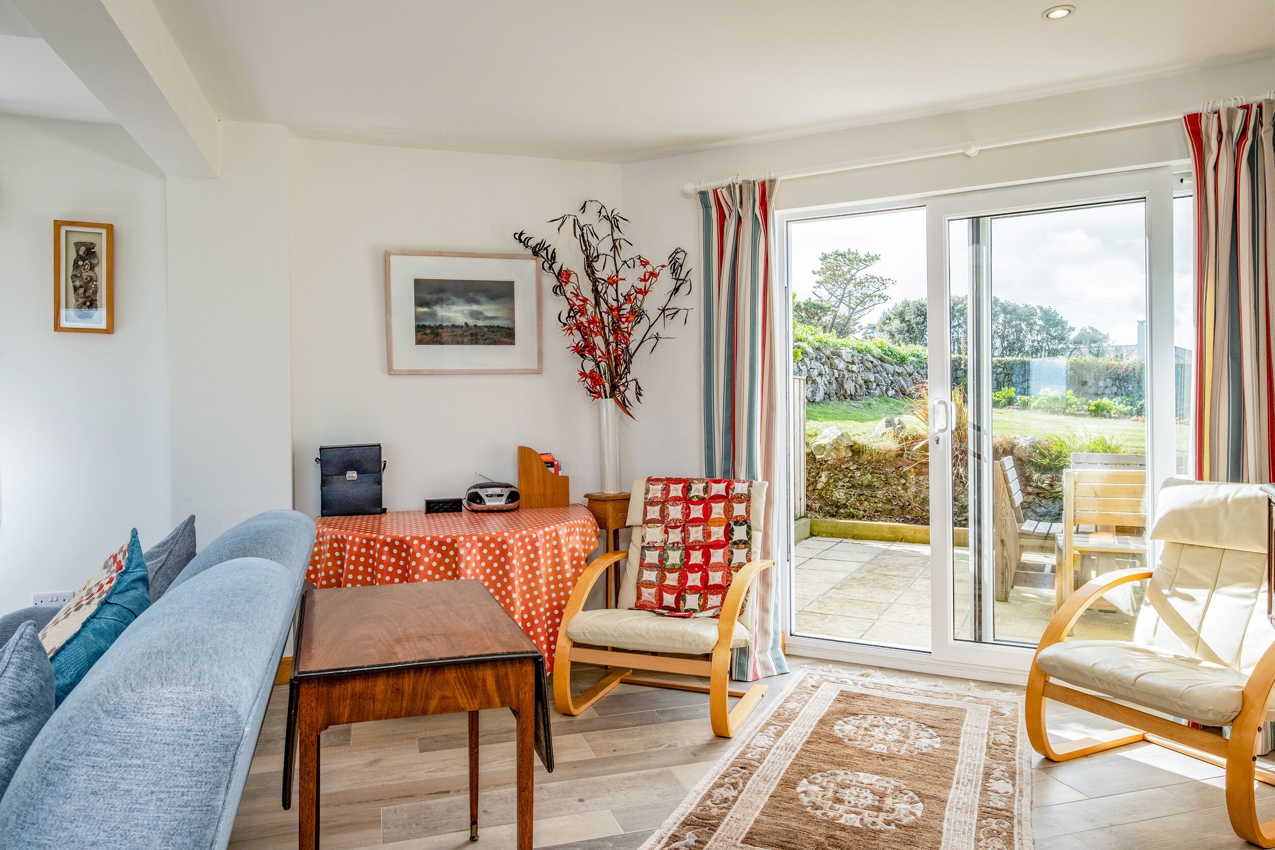Sala de estar con sofá, mesa de madera, una silla y una mesa de comedor con mantel rojo. Hay una ventana grande con cortinas, un cuadro enmarcado en la pared y una planta decorativa.
