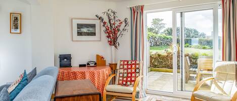 Sala de estar con sofá, mesa de madera, una silla y una mesa de comedor con mantel rojo. Hay una ventana grande con cortinas, un cuadro enmarcado en la pared y una planta decorativa.