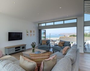A spacious living room with a large TV, comfortable seating, and a view of the beach. - Point Break (Wadebridge)