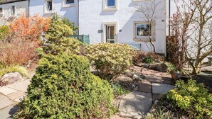 Une petite maison blanche avec un porche, entourée d’un jardin où poussent différentes plantes et bordée d’un chemin en pierre.