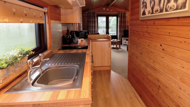 A wooden cabin interior with a kitchen area featuring a sink, stove, and microwave. There is a dining area with a table and chairs, and a window with a view. - Holly Lodge (Saltburn-by-the-Sea)