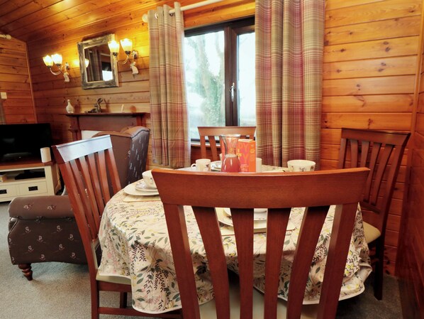 A wooden cabin interior with a dining table set for four, surrounded by wooden chairs. A television is mounted on the wall, and there are two wall-mounted lamps. - Holly Lodge (Saltburn-by-the-Sea)