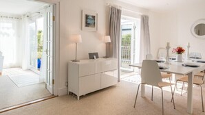 A modern, well-lit room with a dining area, a white cabinet with a picture, and a view of the outdoors. - The Moorings (Wadebridge)