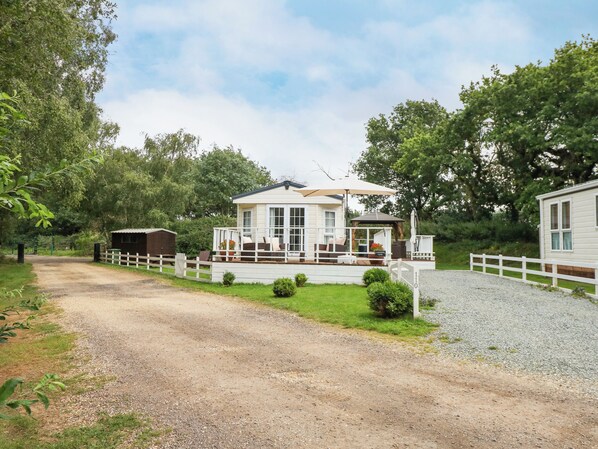 A white mobile home with a porch and outdoor seating area, surrounded by trees and a gravel driveway. - Lakeside View (King's Lynn)