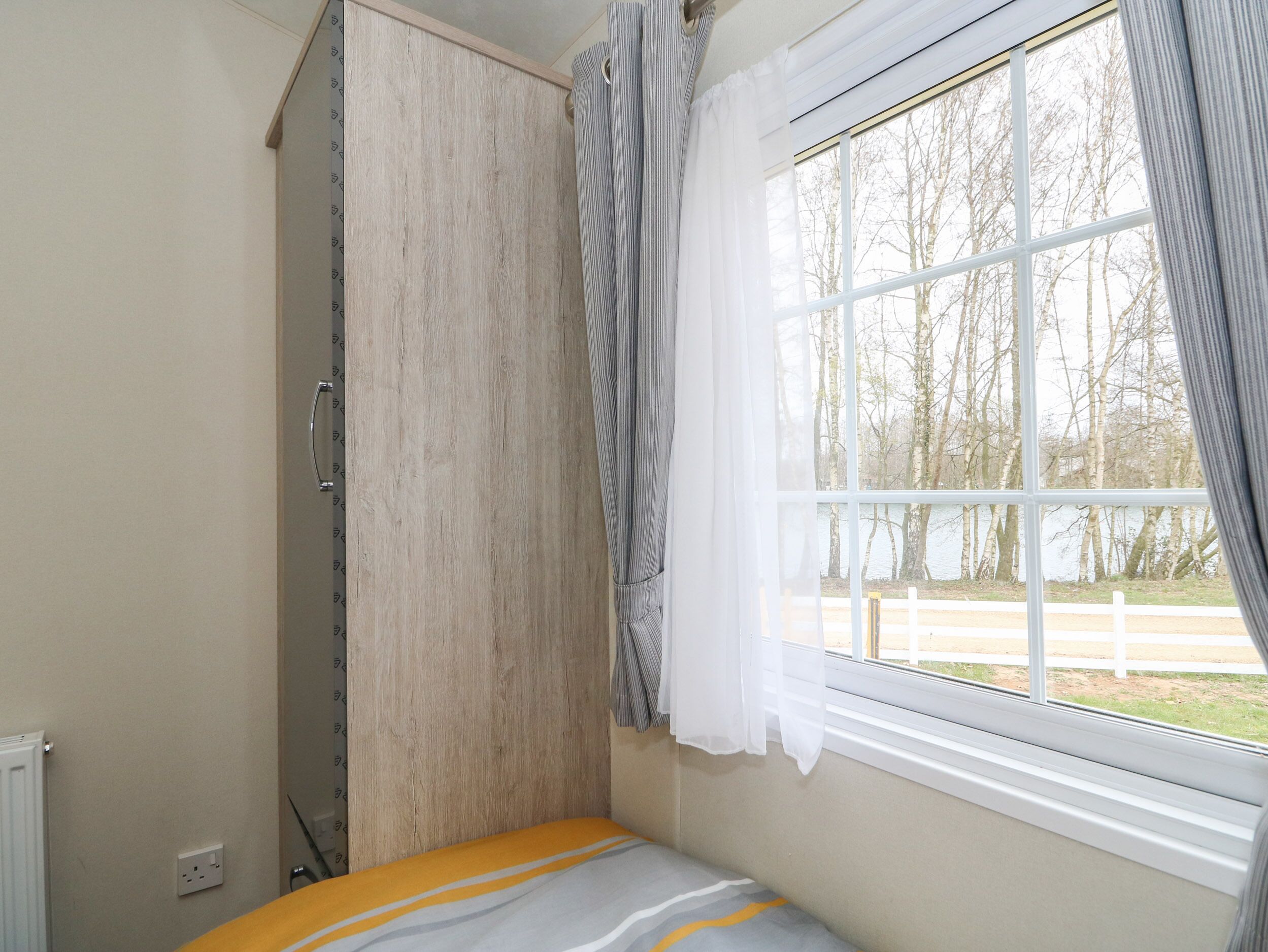 A bedroom with a bed, a wardrobe, a window with curtains, and a view of trees and a fence.
