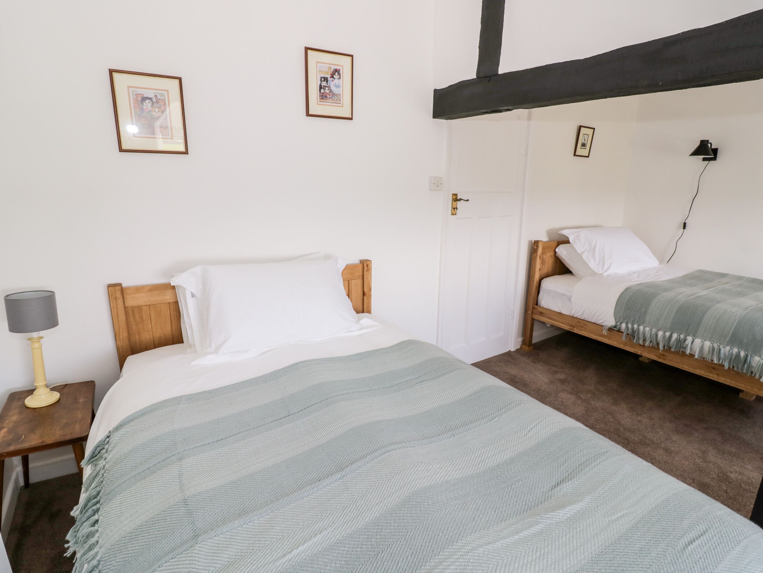 A room with two beds, a wooden headboard, a bedside table with a lamp, and two framed pictures on the wall.