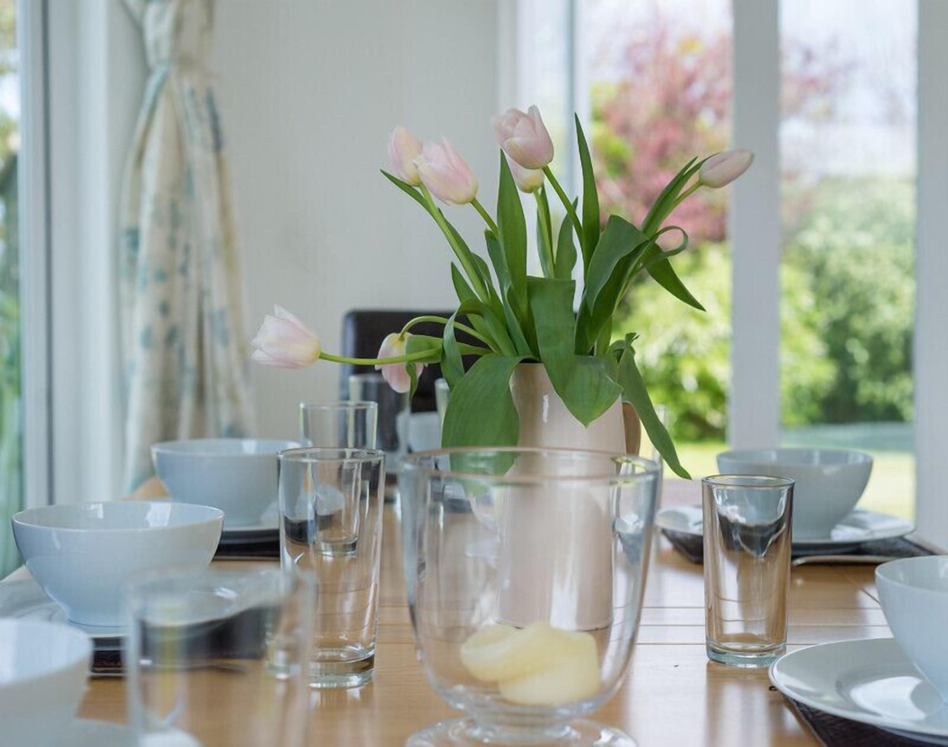 Una mesa de comedor con un jarrón de tulipanes rosados, copas de cristal y un bol de mantequilla.