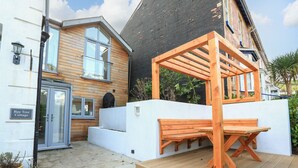 A wooden deck with a bench and a pergola, a white wall, and a black brick building with a window. - Bay Tree Cottage, Dartmouth (Dartmouth)