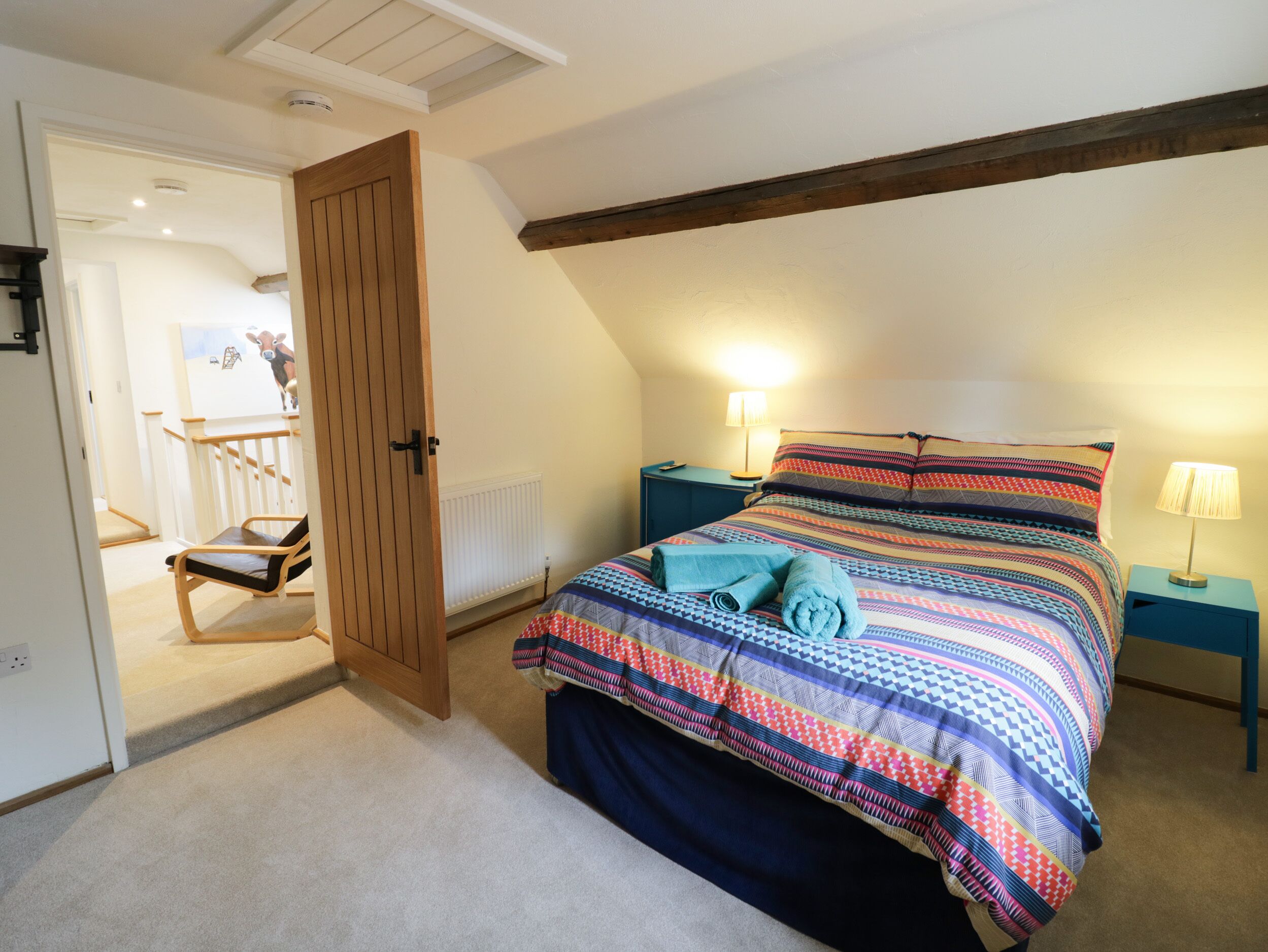 A bedroom with a sloped ceiling, a bed with a colorful quilt, two bedside lamps, and a wooden door leading to another room.