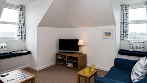 A cozy room with a TV, a small table with books, a lamp, and a blue couch. - Pinewood 14 (Wadebridge)