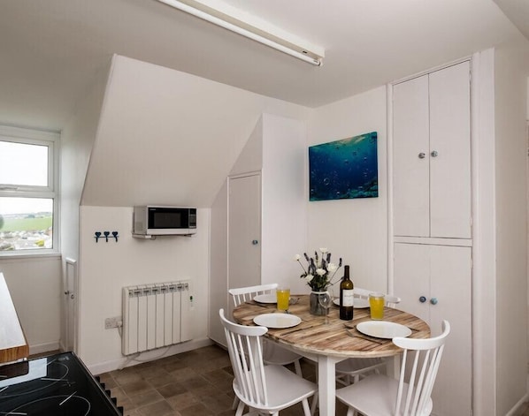 A dining area with a round wooden table, white chairs, a small painting, a white cabinet, and a microwave on the wall.