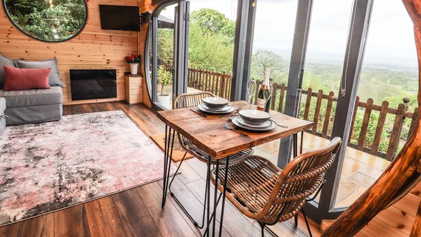 Interior acolhedor de uma cabana com uma mesa de jantar posta para dois, um sofá e uma grande janela com vista panorâmica.