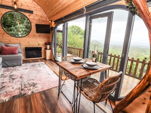 A cozy cabin interior with a dining table set for two, a sofa, and a large window offering a scenic view. - Offas Dyke Escape (Saint Asaph)
