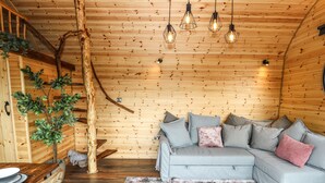 Interior acolhedor de uma cabana, com teto de madeira, sofá cinza com almofadas, mesa de jantar com cadeiras e uma árvore decorativa.